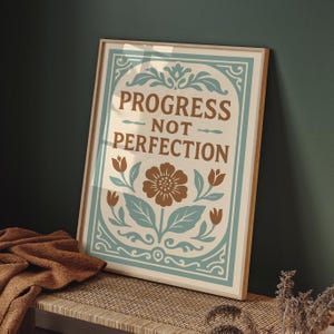 Puede incluir: Un cuadro enmarcado con las palabras "PROGRESS NOT PERFECTION" en letras marrones. El diseño incluye una flor central y detalles florales en marrón y verde azulado sobre un fondo crema. El marco es de color madera clara.