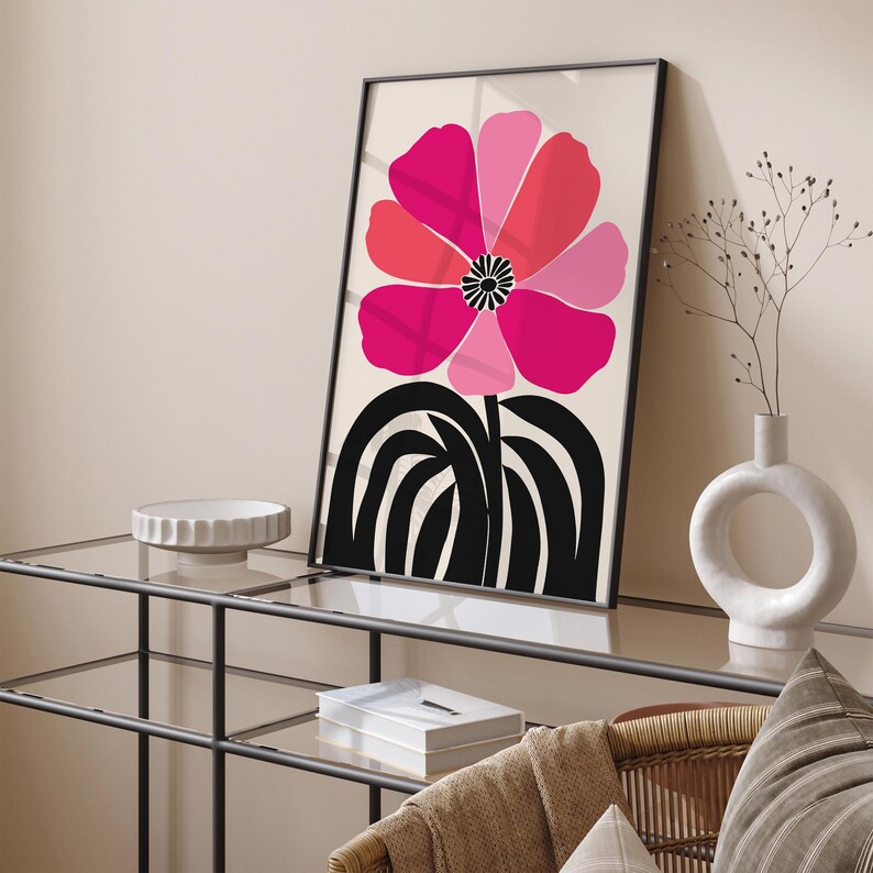 Op de afbeelding: Een ingelijste kunstprint met een gestileerde bloem in roze en rode tinten tegen een cr&egrave;mekleurige achtergrond. De bloem heeft een zwart-wit centrum en een zwarte stengel. Het kunstwerk wordt gepresenteerd op een glazen tafel.
