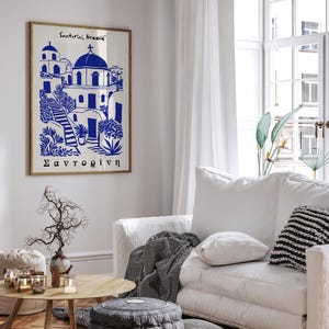 Puede incluir: Una ilustraci&oacute;n azul y blanca de un pueblo griego con el texto "Santorini, Grecia" y "Santorini" en griego. La ilustraci&oacute;n est&aacute; enmarcada y colgada en una pared blanca.