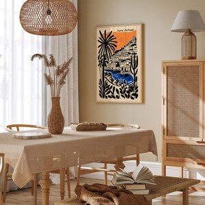 May include: A framed art print featuring a stylized illustration of Porto, Portugal. The artwork showcases a coastal scene with buildings, palm trees, and a blue body of water against an orange sky. The print is displayed in a room with a neutral color palette.