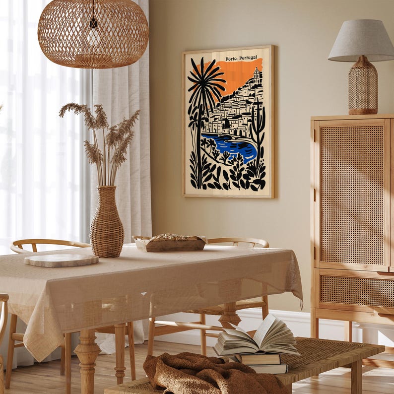 May include: A framed art print featuring a stylized illustration of Porto, Portugal. The artwork showcases a coastal scene with buildings, palm trees, and a blue body of water against an orange sky. The print is displayed in a room with a neutral color palette.