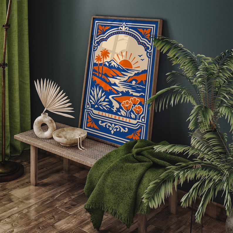 May include: A framed art print featuring a California landscape design in shades of blue, orange, and cream. The artwork depicts a sunset over mountains, palm trees, and poppies, with the word "CALIFORNIA" at the bottom. The print is displayed on a wooden bench.