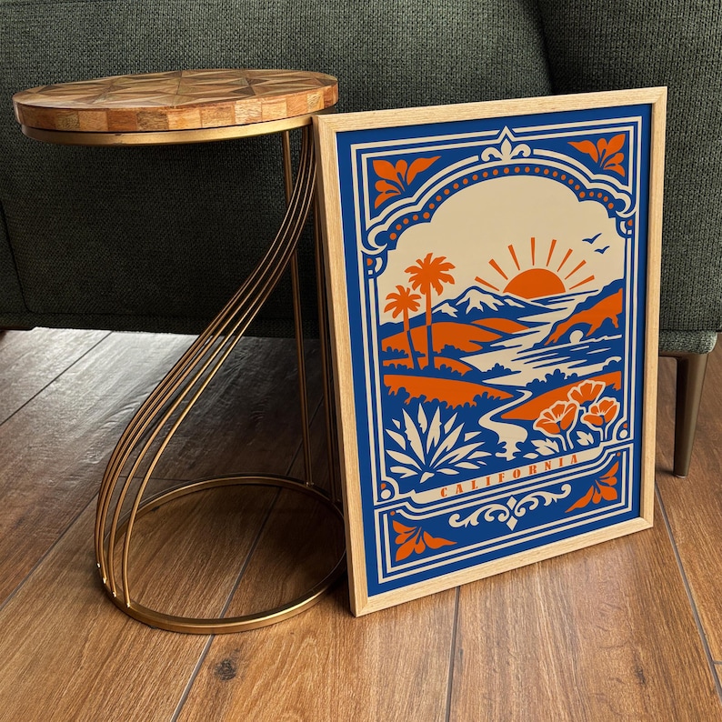 May include: A framed rectangular print featuring a stylized California landscape with orange mountains, palm trees, a sun, and flowers against a blue background. The word CALIFORNIA is printed at the bottom. The print is displayed next to a side table.