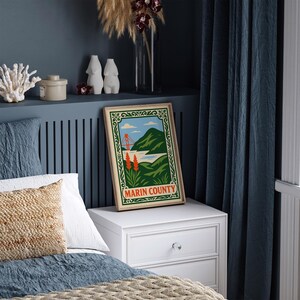 May include: A framed poster of Marin County with a vintage travel design showing the Golden Gate Bridge, green hills, and the text "Marin County." The poster is on a white bedside table next to a bed with blue bedding.