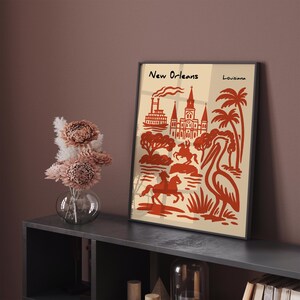 Op de afbeelding: Ingelijste kunstdruk met een oranje illustratie van bezienswaardigheden in New Orleans op een cr&egrave;mekleurige achtergrond. De print bevat de tekst "New Orleans Louisiana". Het kunstwerk wordt tentoongesteld op een donkergrijze plank, met een vaas bloemen.