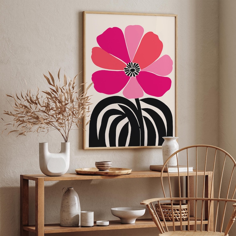 Op de afbeelding: Een ingelijste kunstdruk met een grote, gestileerde bloem in roze en rode tinten tegen een cr&egrave;mekleurige achtergrond. De bloem heeft een zwart hart en staat tegen zwarte, gebogen lijnen. Het kunstwerk wordt getoond in een kamer met houten meubels.