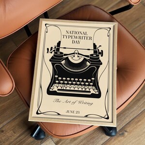 May include: A rectangular framed print featuring a black illustration of a vintage typewriter on a cream background. The text reads NATIONAL TYPEWRITER DAY, The Art of Writing, and JUNE 23. The design includes decorative black border lines.