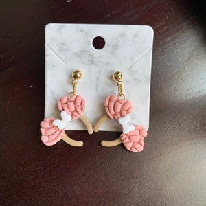 May include: A pair of pink and white dangle earrings with a gold-tone post. The earrings are shaped like a concha, a traditional Mexican sweet bread.