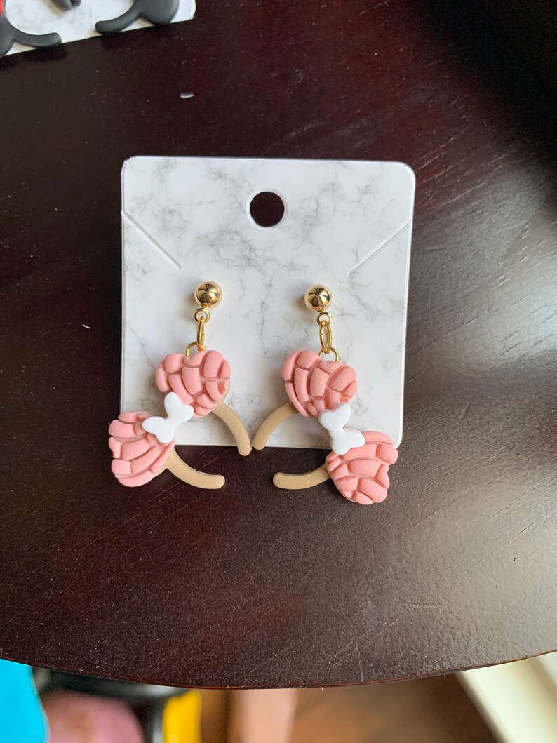 May include: A pair of pink and white dangle earrings with a gold-tone post. The earrings are shaped like a concha, a traditional Mexican sweet bread.