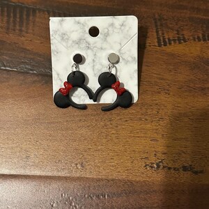 May include: A pair of black Mickey Mouse ears with red bows, displayed on a white marble background. The earrings are made of polymer clay and have silver hooks.