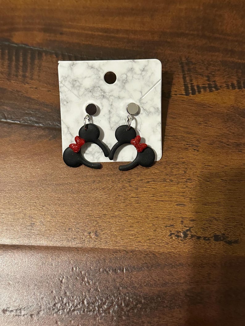 May include: A pair of black Mickey Mouse ears with red bows, displayed on a white marble background. The earrings are made of polymer clay and have silver hooks.