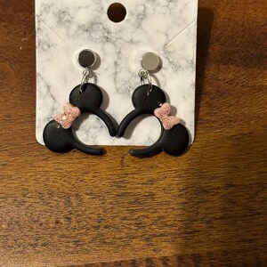May include: A pair of black dangle earrings with pink glitter bows. The earrings are shaped like Mickey Mouse ears.