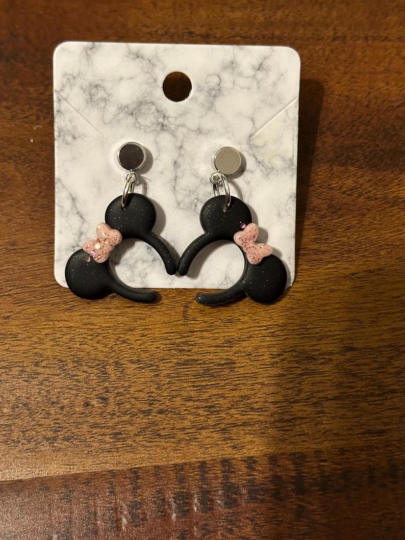 May include: A pair of black dangle earrings with pink glitter bows. The earrings are shaped like Mickey Mouse ears.