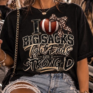 Könnte beinhalten: Ein schwarzes T-Shirt mit einer Grafik aus einem Football-Herz, einer Schleife mit Leopardenmuster und dem Text I LOVE BIG SACKS TIGHT ENDS AND A STRONG D. Dazu gehört ein weißer Becher, der mit braunen Footballs und Leoparden-Schleifen verziert ist.