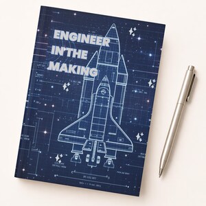 May include: A navy blue notebook with a blueprint-style illustration of a space shuttle and the text "ENGINEER IN THE MAKING." A silver pen rests beside the notebook on a white surface.