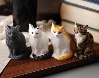 Könnte beinhalten: Vier Keramik-Katzenfiguren, jede mit einer anderen Farbe und einem anderen Muster. Die Katzen sitzen auf einer Holzoberfläche. Die Katzen sind schwarz, weiß, orange und braun.