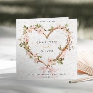Puede incluir: Una tarjeta de felicitación cuadrada con una corona en forma de corazón hecha de flores rosas y ramas marrones. El texto dice CONGRATULATIONS, CHARLOTTE and OLIVER, ON YOUR 25TH SILVER WEDDING ANNIVERSARY, 20.09.2026. Un lazo rosa está atado abajo.