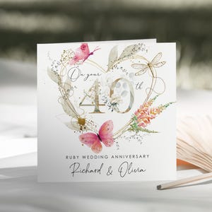 May include: A square greeting card featuring a gold heart outline, pink butterflies, a dragonfly, and floral accents. The text reads On your 40th, RUBY WEDDING ANNIVERSARY, and Richard & Olivia. The design is printed on a white background with gold details.