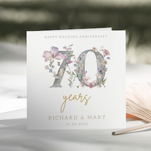 Puede incluir: Una tarjeta blanca con un diseño floral en tonos morados y rosas. El número 70 está rodeado de flores y el texto "Happy Wedding Anniversary" está en la parte superior. El texto "years" está escrito en cursiva dorada debajo del número. Los nombres "Richard & Mary" y la fecha "01.05.2025" están impresos en la parte inferior de la tarjeta.