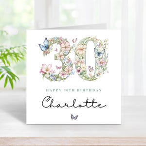 Puede incluir: Una tarjeta de felicitación cuadrada con el número 30 formado por ilustraciones en acuarela de flores rosas, blancas y azules con mariposas azules. Debajo del número, el texto dice HAPPY 30TH BIRTHDAY y el nombre Charlotte en cursiva.
