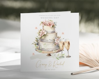 May include: A white wedding cake with pink flowers and green leaves, decorated with a gray ribbon. Two champagne flutes are on a table next to the cake. The card reads "Congratulations on your Wedding Day Gary & Rachel 20.06.2025".