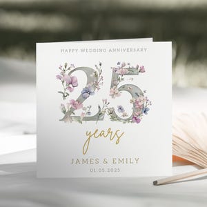 Peut inclure: Une carte blanche avec un motif floral autour du chiffre 25. Le texte "Happy Wedding Anniversary" est en haut de la carte. Le texte "25 years" est sous le chiffre. Les noms "James & Emily" et la date "01.05.2025" sont en bas de la carte.