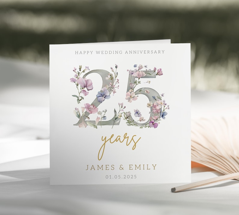Peut inclure: Une carte blanche avec un motif floral autour du chiffre 25. Le texte "Happy Wedding Anniversary" est en haut de la carte. Le texte "25 years" est sous le chiffre. Les noms "James & Emily" et la date "01.05.2025" sont en bas de la carte.