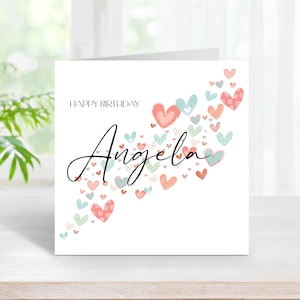 May include: A white card with the text "Happy Birthday" and the name "Angela" written in cursive. The name is surrounded by a scattering of pink and blue hearts with different patterns.