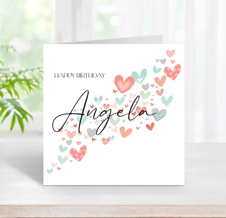 May include: A white card with the text "Happy Birthday" and the name "Angela" written in cursive. The name is surrounded by a scattering of pink and blue hearts with different patterns.