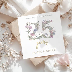 Peut inclure: Une carte blanche avec une inscription dor&eacute;e qui dit "Happy Wedding Anniversary 25 years James & Emily". Le nombre 25 est fait de gris avec des fleurs violettes et roses autour.
