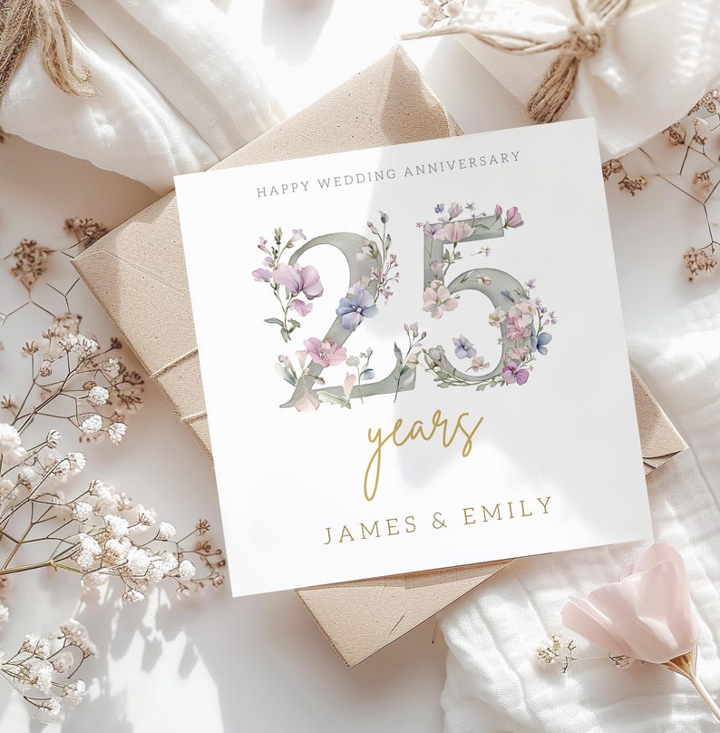 Peut inclure: Une carte blanche avec une inscription dor&eacute;e qui dit "Happy Wedding Anniversary 25 years James & Emily". Le nombre 25 est fait de gris avec des fleurs violettes et roses autour.