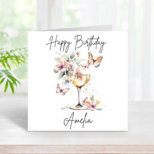 Puede incluir: Una tarjeta blanca con el texto "Happy Birthday Amelia" en letra cursiva negra. Una ilustración de acuarela de una copa de vino con flores y mariposas está en el centro de la tarjeta.