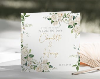 May include: White wedding card with a floral design featuring white roses and green leaves. The card reads "With love on your wedding day Charlotte & Henry 20.06.2025".
