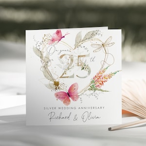 Puede incluir: Una tarjeta de felicitaci&oacute;n cuadrada para un 25 aniversario de bodas con un contorno de coraz&oacute;n dorado, mariposas rosas, una lib&eacute;lula y detalles florales. El texto dice On your 25th, SILVER WEDDING ANNIVERSARY y los nombres Richard & Olivia.