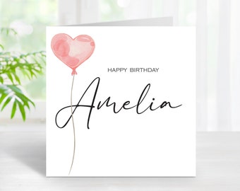 Peut inclure: Une carte de vœux blanche avec un ballon en forme de cœur rose à l'aquarelle. La carte indique "Happy Birthday Amelia" en écriture noire. Le ballon est attaché à une fine ficelle marron.