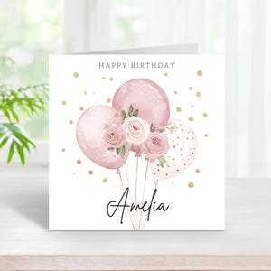 May include: A square birthday card with a watercolor design. The card features pink balloons, roses, and the name "Amelia" in cursive. The words "Happy Birthday" are at the top, with gold confetti dots scattered around.