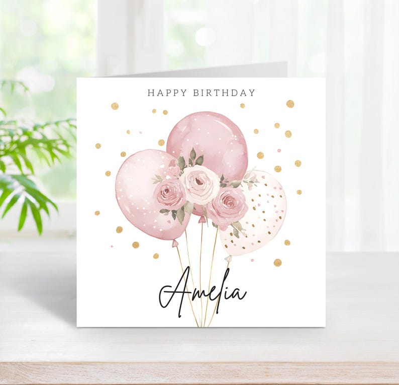 May include: A square birthday card with a watercolor design. The card features pink balloons, roses, and the name "Amelia" in cursive. The words "Happy Birthday" are at the top, with gold confetti dots scattered around.