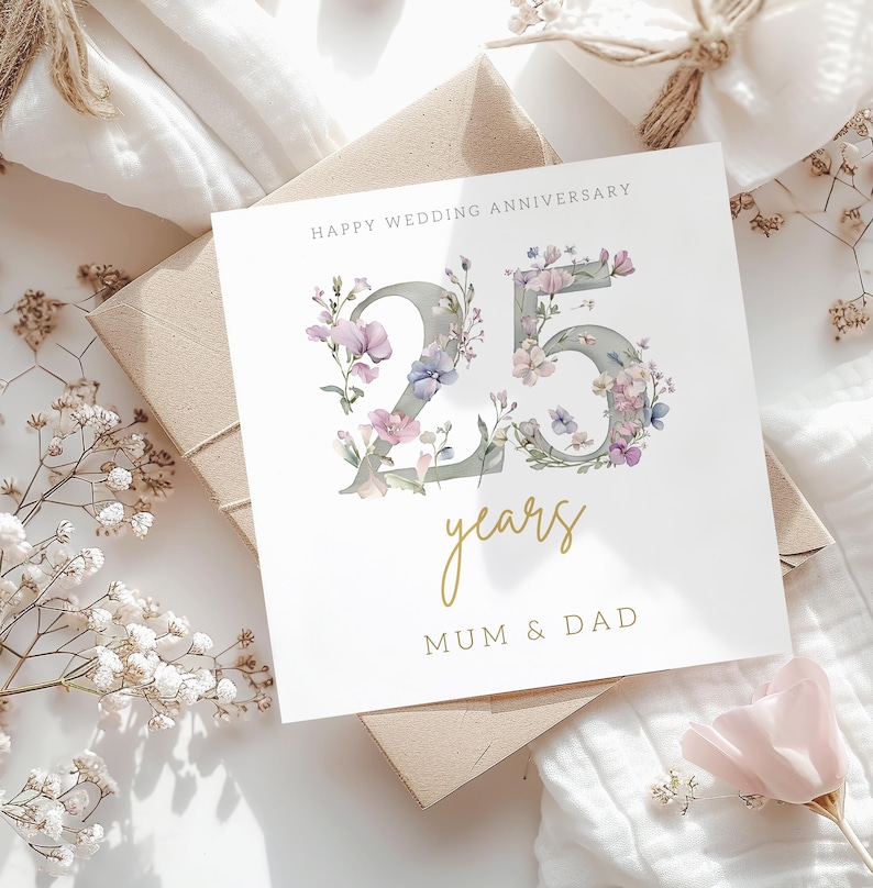 Peut inclure: Une carte blanche avec une inscription dor&eacute;e qui dit "Happy Wedding Anniversary 25 years Mum & Dad". Le nombre 25 est fait de gris et est d&eacute;cor&eacute; de fleurs violettes et roses.