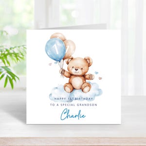 May include: A square greeting card featuring a watercolor illustration of a brown teddy bear sitting on a blue cloud, holding two balloons, one blue with the number 1 and one beige. Text reads: HAPPY 1ST BIRTHDAY TO A SPECIAL GRANDSON Charlie.