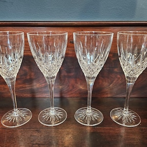 May include: Four clear crystal wine glasses with a diamond-cut pattern on the bowl and a starburst design on the base. The glasses are displayed on a dark wooden surface.