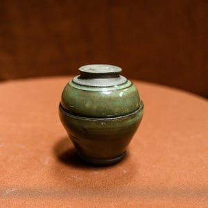 May include: A small, handcrafted ceramic jar with a lid. The jar is a deep green colour with lighter green and grey accents around the top. The jar is sitting on a brown surface.