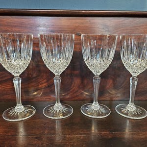 May include: Four clear crystal wine glasses with a diamond pattern on the bowl and vertical cuts. The glasses have a long stem and a round base with a starburst design. The glasses are on a dark wooden surface.