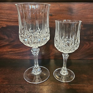 May include: Two clear crystal wine glasses with intricate diamond and vertical line patterns. The glasses have a flared bowl, a detailed stem, and a round base. The glasses are displayed on a dark wooden surface.
