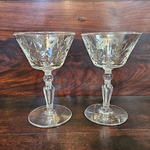 Puede incluir: Dos copas de champán transparentes y vintage con diseños florales grabados. Las copas tienen un cuenco ancho, un tallo decorativo y una base circular. El fondo es una superficie de madera oscura.