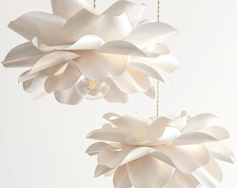 May include: Two off-white pendant lights shaped like blooming flowers with layered, curved petals. Each light fixture hangs from a thin, twisted cord, featuring a central exposed light bulb nestled within the soft, organic petal structure.