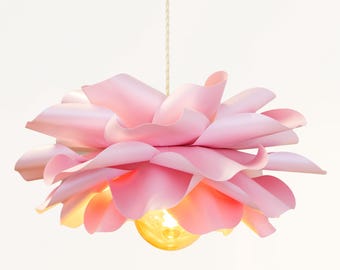 May include: A hanging pendant light fixture featuring a layered, petal-like shade in soft shades of pink. The structure mimics the appearance of a blooming flower, with a single warm-toned light bulb visible at the center, suspended by a cord.