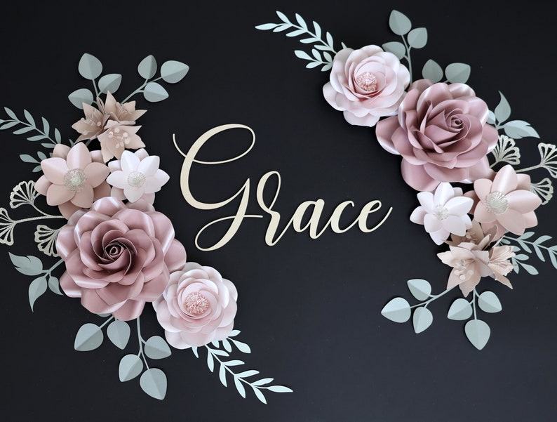 May include: A black background with a paper flower arrangement in shades of pink and white. The flowers are arranged in a semi-circle with the word "Grace" written in cursive in the center.