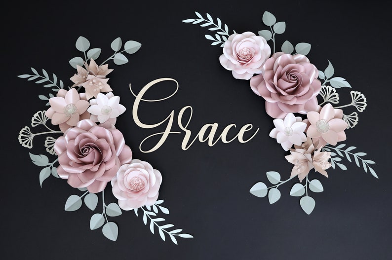 May include: A paper flower arrangement with the word "Grace" in the center. The flowers are in shades of pink and white, with green leaves. The background is black.