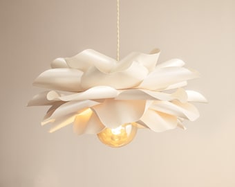 May include: A white pendant light fixture featuring layered, curved, petal-like shades that form a flower-like shape. A single warm-toned light bulb is visible at the center, suspended from a thin, textured cord against a plain background.