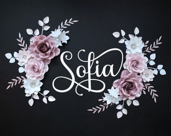 May include: A paper flower arrangement with the name "Sofia" written in white script. The flowers are pink and white and have a delicate, layered design. The arrangement is perfect for a baby shower or nursery decor.